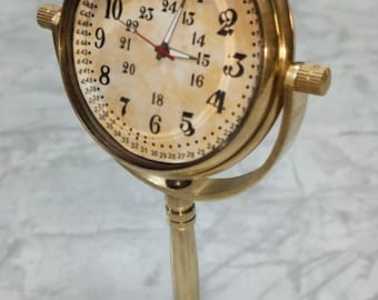 May include: A brass-colored desk clock with a circular face featuring black Arabic numerals from 1 to 12, an inner ring of numbers from 13 to 24, and a minute track from 25 to 60. The clock is mounted on a vertical stand with a round, engraved base.