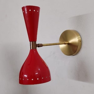 May include: A red, mid-century modern wall sconce with a brass arm and mounting plate. The sconce has a conical shade with small white dots and a flared top. The fixture is mounted on a white wall.