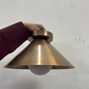 May include: A bronze ceiling light fitting with a conical shade and a round, white globe. The fitting has a circular base. The light fixture is designed for indoor use. The fitting is held by a person's hand.