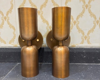 May include: Two matching bronze-colored wall sconces. Each sconce has a cylindrical base and a flared, cup-shaped top. The sconces are mounted on circular backplates. The background features a patterned wallpaper.