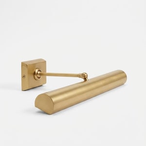 May include: A brass-toned picture light with a rectangular wall mount and a cylindrical light fixture. The light has a pivoting arm and a rounded end. The fixture is designed to illuminate artwork or other wall-mounted items.