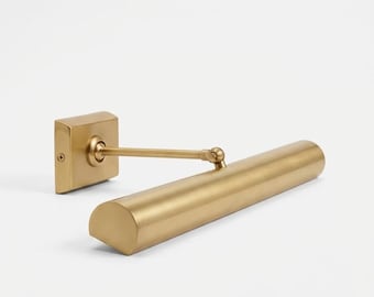May include: A brass-toned picture light with a rectangular wall mount and a cylindrical light fixture. The light has a pivoting arm and a rounded end. The fixture is designed to illuminate artwork or other wall-mounted items.