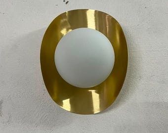 May include: A wall-mounted light fixture featuring a circular, brushed gold metal backplate with a curved, reflective surface. Centered on the gold plate is a spherical, frosted white glass globe that serves as the light diffuser.