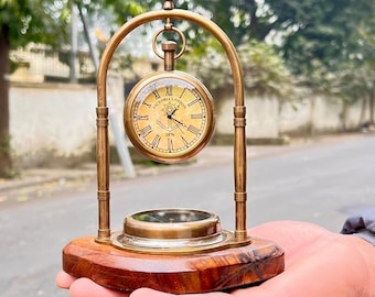 May include: A vintage-style brass clock with a cream-colored face and Roman numerals. The clock is suspended within a brass arch and mounted on a wooden base. The clock face reads "VICTORIA LONDON 1876".