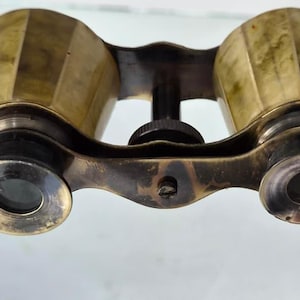 May include: Vintage brass opera glasses featuring a metallic gold finish with a faceted barrel design. The optical device includes two circular eyepieces, a central focus adjustment knob, and a thin, extendable metal handle attached to the side.