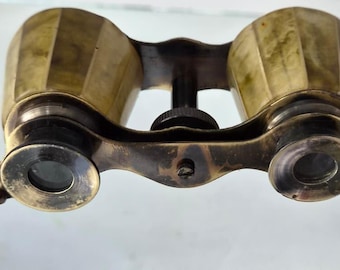 May include: Vintage brass opera glasses featuring a metallic gold finish with a faceted barrel design. The optical device includes two circular eyepieces, a central focus adjustment knob, and a thin, extendable metal handle attached to the side.