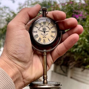 Op de afbeelding: Een vintage zakhorloge met een standaard. De wijzerplaat is crèmekleurig met zwarte Romeinse cijfers en de woorden "VICTORIA LONDON 1876". Het horloge wordt vastgehouden door een messing standaard met een cirkelvormige basis. Het horloge heeft een diameter van ongeveer 7,6 cm.