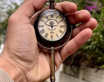 May include: A vintage pocket watch with a stand. The watch face is cream-colored with black Roman numerals and the words "VICTORIA LONDON 1876". The watch is held by a brass stand with a circular base. The watch is approximately 3 inches in diameter.