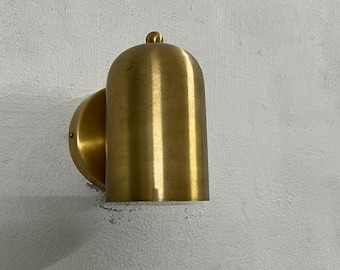 May include: A wall-mounted light fixture featuring a cylindrical brass body with a rounded top and a small spherical finial. The metal has a brushed finish, and the fixture is attached to a circular brass base plate.