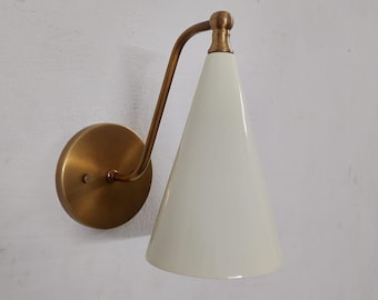 May include: A wall-mounted sconce with a brass finish and a white, cone-shaped shade. The sconce features a circular base and a curved arm connecting to the shade. The design is minimalist and modern, suitable for interior lighting.