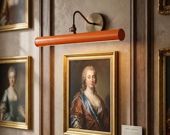 May include: An orange art gallery light fixture with a bronze-colored arm and mounting plate is mounted above a framed portrait. The light illuminates the artwork. Other framed portraits are visible on the wall.