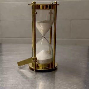 May include: A gold-coloured hourglass with a clear glass centre filled with white sand. The hourglass has a decorative, ornate design with a base and top, and a small metal lever. The sand is flowing from the top to the bottom.