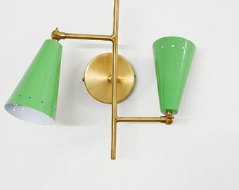 May include: A wall-mounted light fixture featuring a central brass-toned vertical rod and a circular mounting plate. Two adjustable arms extend from the rod, each holding a conical, light green metal shade with small perforations.