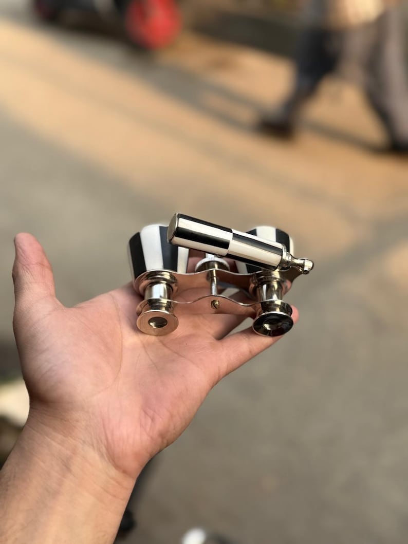 May include: A pair of opera glasses with a black and white striped design on the body and a silver-colored handle and trim. The opera glasses are held in a hand.