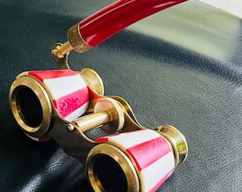 May include: Vintage opera glasses with a red handle and red and white striped barrels. The brass-colored metal frame holds the lenses and the handle. The opera glasses are set against a dark background.