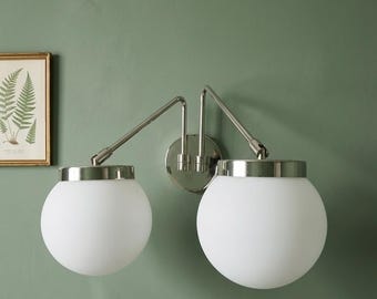 May include: A modern wall sconce with two white globe shades and a brushed nickel finish. The fixture has adjustable arms and is mounted on a round backplate. The sconce is mounted on a green wall.