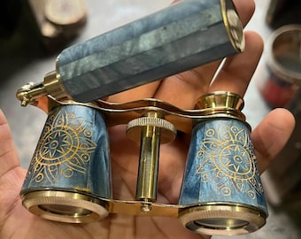 May include: A pair of opera glasses featuring a polished brass frame and blue marbled barrels. The barrels are decorated with intricate gold-colored sunburst engravings. A central brass focus wheel sits between the two eyepieces, and a handle is attached.