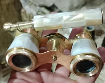 May include: Antique opera glasses with a gold-tone metal frame and white mother-of-pearl accents. The binoculars have a handle with a matching mother-of-pearl finish. The lenses are black. A vintage item.