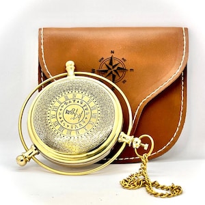 May include: A gold-colored compass with Roman numerals and intricate details, displayed with a brown leather case. The case features a compass rose design. A gold chain is attached to the compass.