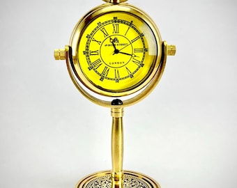 May include: A gold-colored desk clock with a yellow face and black Roman numerals. The clock has a stand with an ornate base and the text "49 Bond Street LONDON". The clock is approximately 5 inches tall.