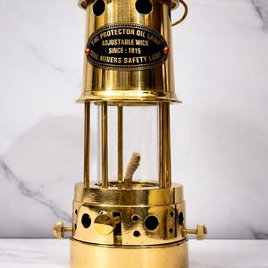 Peut inclure: Une lampe à huile vintage en laiton avec un cylindre en verre transparent et une mèche réglable. La lampe porte l'inscription "THE PROTECTOR OIL LAMP ADJUSTABLE WICK SINCE: 1815 COAL MINERS SAFETY LAMP" sur une étiquette ovale noire.