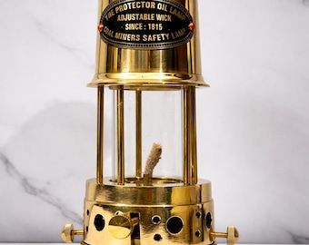 May include: A vintage brass oil lamp with a clear glass cylinder and adjustable wick. The lamp features the text "THE PROTECTOR OIL LAMP ADJUSTABLE WICK SINCE: 1815 COAL MINERS SAFETY LAMP" on a black oval label. The lamp has a handle and several air holes.