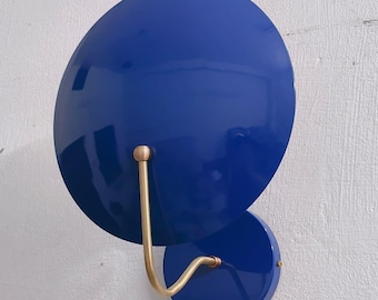 May include: A modern wall sconce featuring two overlapping, glossy, royal blue circular shades. A curved, brass-colored metal arm connects the shades. The sconce is mounted on a textured, off-white wall. This decorative lighting fixture adds a pop of color.