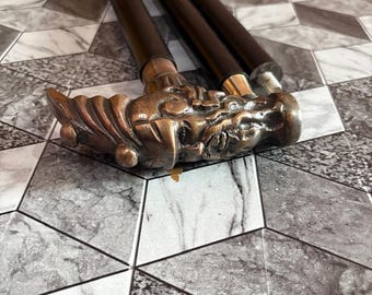 May include: A decorative walking cane with a bronze dragon head handle and a dark brown wooden shaft. The cane is lying on a gray and white patterned surface. The dragon head is intricately detailed.