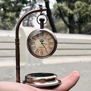 May include: A bronze-coloured pocket watch with a cream face, Roman numerals, and the words "VICTORIA LONDON 1915." The watch hangs from a curved stand with a circular base. The watch is approximately 5 cm in diameter.