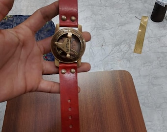 May include: A vintage wristwatch with a brass face and a red leather strap. The watch face has a compass design with the word "RUT" visible. The strap is secured with a brass buckle and rivets. The strap length is approximately 25 cm.