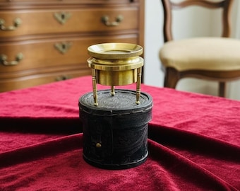 May include: A vintage brass magnifying glass with a leather base. The magnifying glass has a circular lens and is supported by three small legs. The base is black and cylindrical, with an embossed design. The item is set on a red velvet surface.