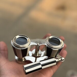 May include: Vintage opera glasses with a black and white checkered pattern on the handle and the sides. The binoculars have a silver-colored metal frame and lenses. The design is classic and elegant.