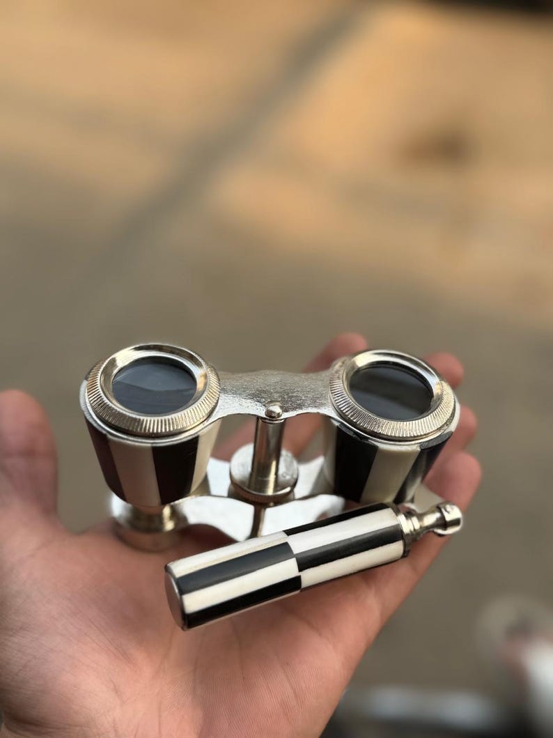 May include: Vintage opera glasses with a black and white checkered pattern on the handle and the sides. The binoculars have a silver-colored metal frame and lenses. The design is classic and elegant.