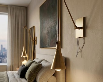 May include: A bedroom interior with a bed, pillows, and a brass wall lamp. The lamp has a cone-shaped shade and an adjustable arm. A painting hangs above the bed, and a window with curtains is in the background.