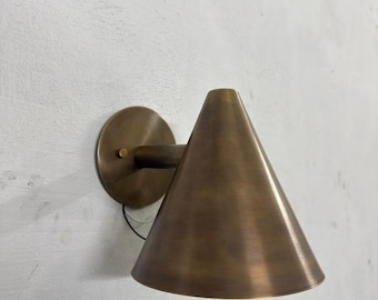 May include: A bronze-colored wall sconce with a conical shade. The sconce has a circular base and a short arm connecting to the shade. The shade is cone-shaped, with a smooth, metallic finish. The sconce is mounted on a white wall.