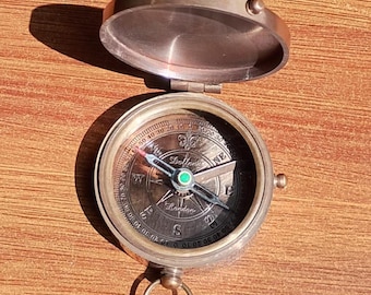 May include: A vintage brass compass with an open lid, revealing a detailed dial with directional markings and a needle. The compass has a small ring for attachment and is set against a wooden surface. The dial reads "London".