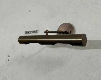 May include: A wall-mounted picture light with a cylindrical bronze-toned metal shade and a semi-circular wall plate. The fixture features an adjustable arm with a pivot joint, connecting the horizontal light housing to the circular mounting base.