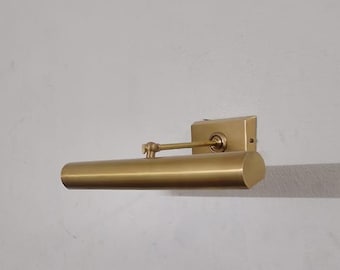 May include: A brass-coloured wall-mounted picture light. The light has a rectangular base and a cylindrical lamp head. The adjustable arm connects the two parts. The fixture is mounted on a light grey wall.