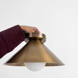 May include: A brass-colored ceiling light fixture with a conical shade and a white, spherical light bulb. The fixture is held against a white background. The shade has a smooth, metallic finish.