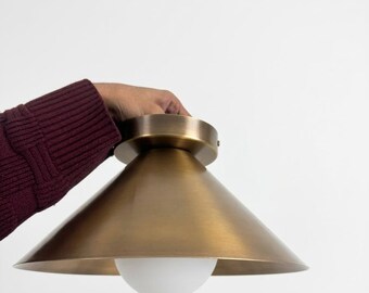 May include: A brass-colored ceiling light fixture with a conical shade and a white, spherical light bulb. The fixture is held against a white background. The shade has a smooth, metallic finish.