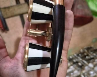 May include: A pair of opera glasses featuring black and white striped barrels with brass accents and a dark brown handle. The device includes a central brass focusing knob and a top-mounted adjustment mechanism for viewing performance events.