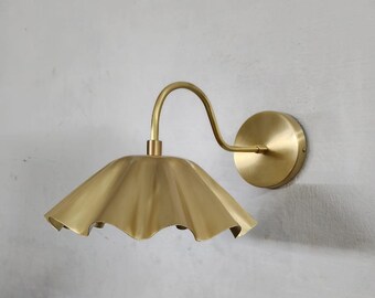 May include: A brass wall sconce with a ruffled shade. The fixture has a curved arm and a circular wall mount. The shade has a scalloped edge, and the overall design is vintage-inspired.