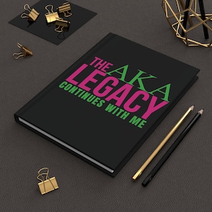 May include: A black journal with the text "THE AKA LEGACY CONTINUES WITH ME" in pink and green. Gold and black pencils and gold binder clips are arranged around the journal on a dark gray surface.