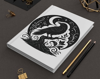 Op de afbeelding: Witte hardcover journal met een zwart ravenontwerp op een tak. Het ontwerp bevat een maan, sterren en decoratief blad. Gouden en zwarte potloden en gouden paperclips zijn ook zichtbaar.