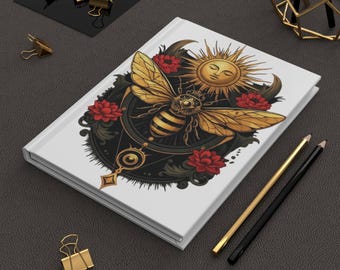May include: White journal with a detailed illustration of a golden bee, sun, and red flowers. The bee is the central focus, with intricate wing and body details. Gold and black pencils and binder clips are also present.
