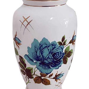 Op de afbeelding: Witte keramische vaas met een gouden rand en een blauw en groen bloemenontwerp. De vaas heeft een grote blauwe roos met groene bladeren en gouden accenten.