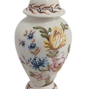 Op de afbeelding: Een witte keramische pot met een deksel, versierd met handgeschilderde bloemenmotieven in tinten blauw, geel, roze en groen.