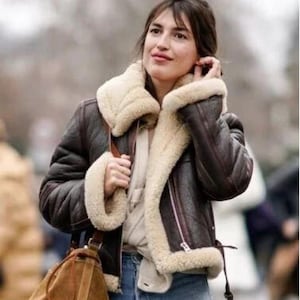 Puede incluir: Chaqueta de cuero marrón con forro y cuello de borreguillo color crema. La chaqueta tiene cierre de cremallera y cintura con hebilla. También se ve un bolso de hombro de ante marrón, junto con unos vaqueros azules.