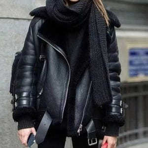 Può includere: Giacca in pelle nera con fodera in montone e sciarpa nera. La giacca presenta una chiusura a zip, cinturini con fibbia sulle maniche e un ampio colletto. Uno zaino nero è visibile sul lato sinistro.