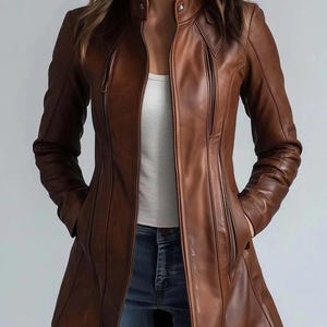 Puede incluir: Chaqueta de cuero marrón con cremallera, mangas largas y cuello alzado. La chaqueta tiene dos bolsillos verticales con cremallera y se lleva sobre una camiseta blanca y vaqueros azules. La chaqueta llega a la rodilla.